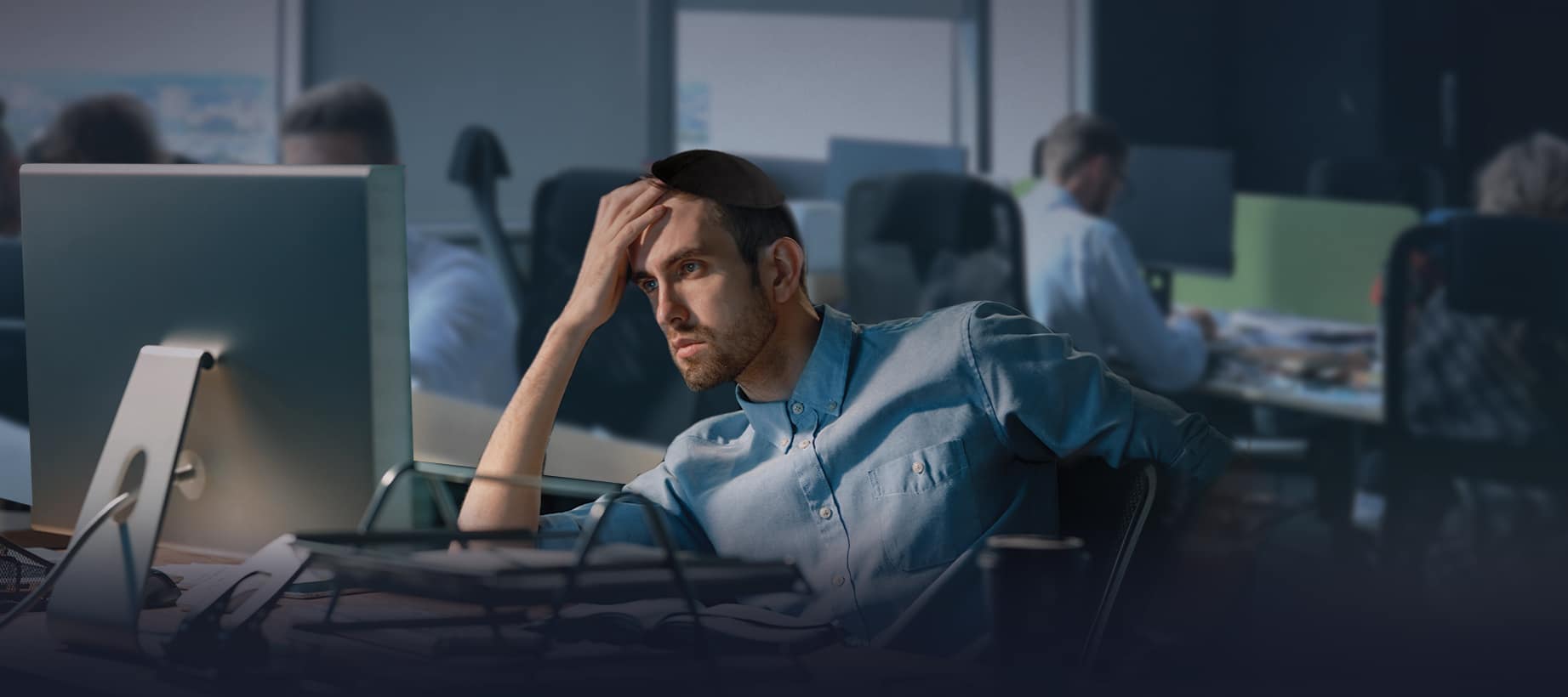 An open-plan office late in the day, lit by monitors, with someone sitting back from a screen.